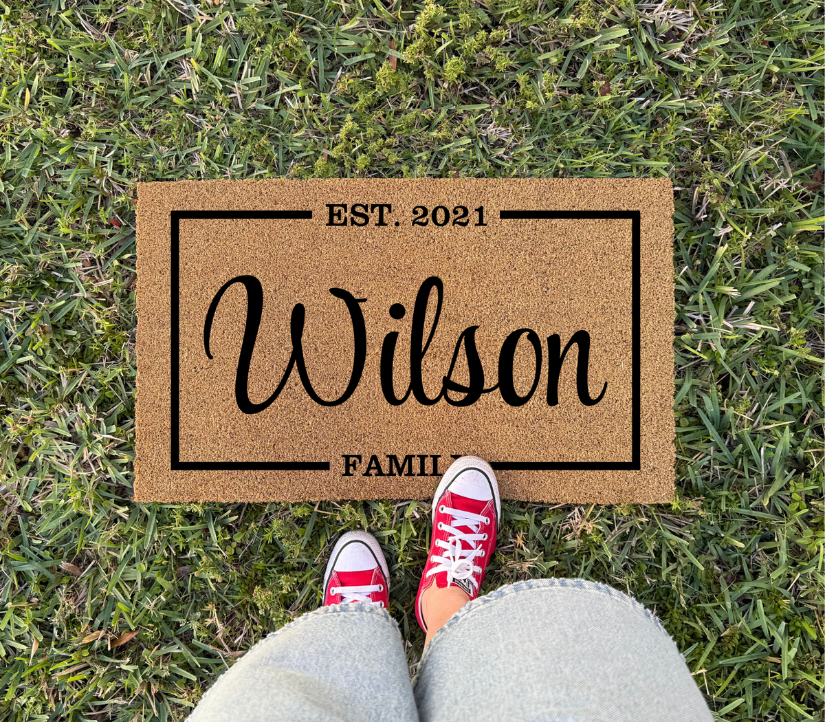 Family name special date doormat with Wilson, EST. 2021 and FAMILY inside a frame, on grass with red sneakers