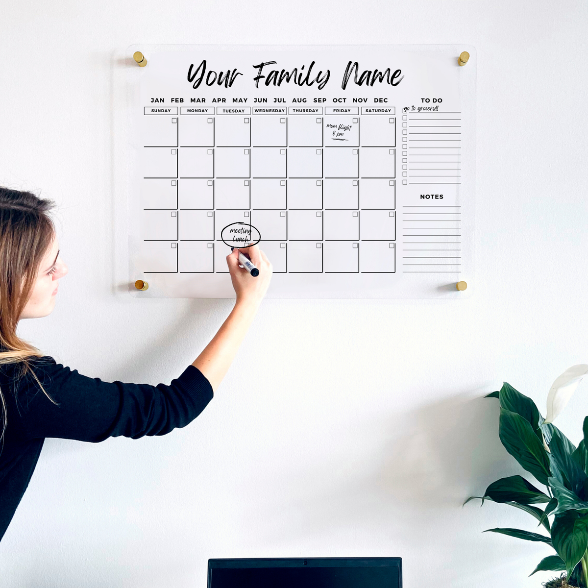 Woman writing on a clear acrylic monthly wall calendar with two side sections above a home office desk.