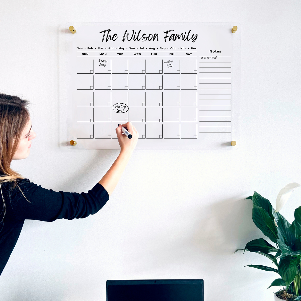 Woman writing on an acrylic monthly calendar with a long notes column above a home office desk.