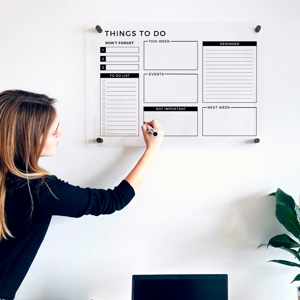 Woman writing on a Things To Do acrylic wall planner with separate lists and reminder boxes above a home office desk.