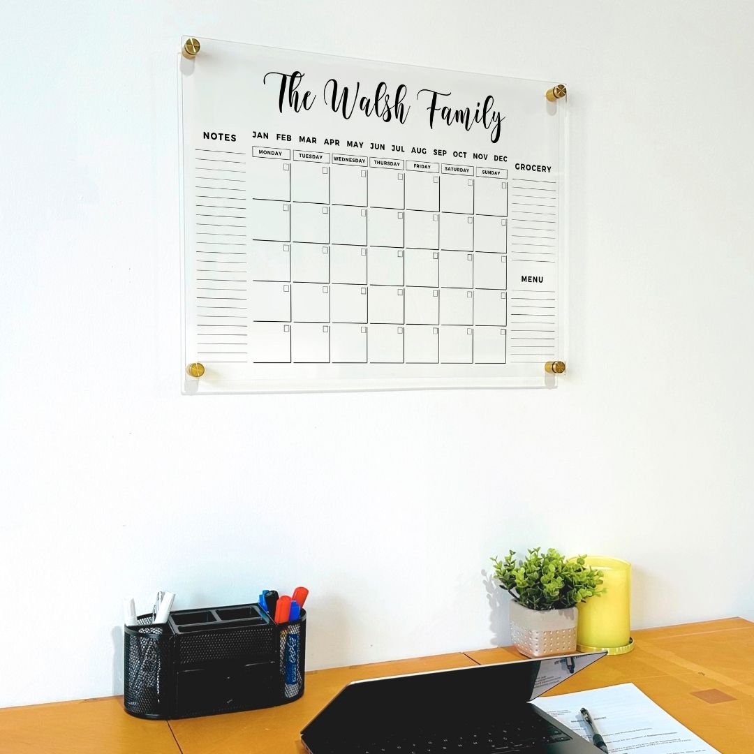 Personalized acrylic family calendar with three side sections and gold mounts, displayed above a wooden desk with a laptop and stationery organizer.