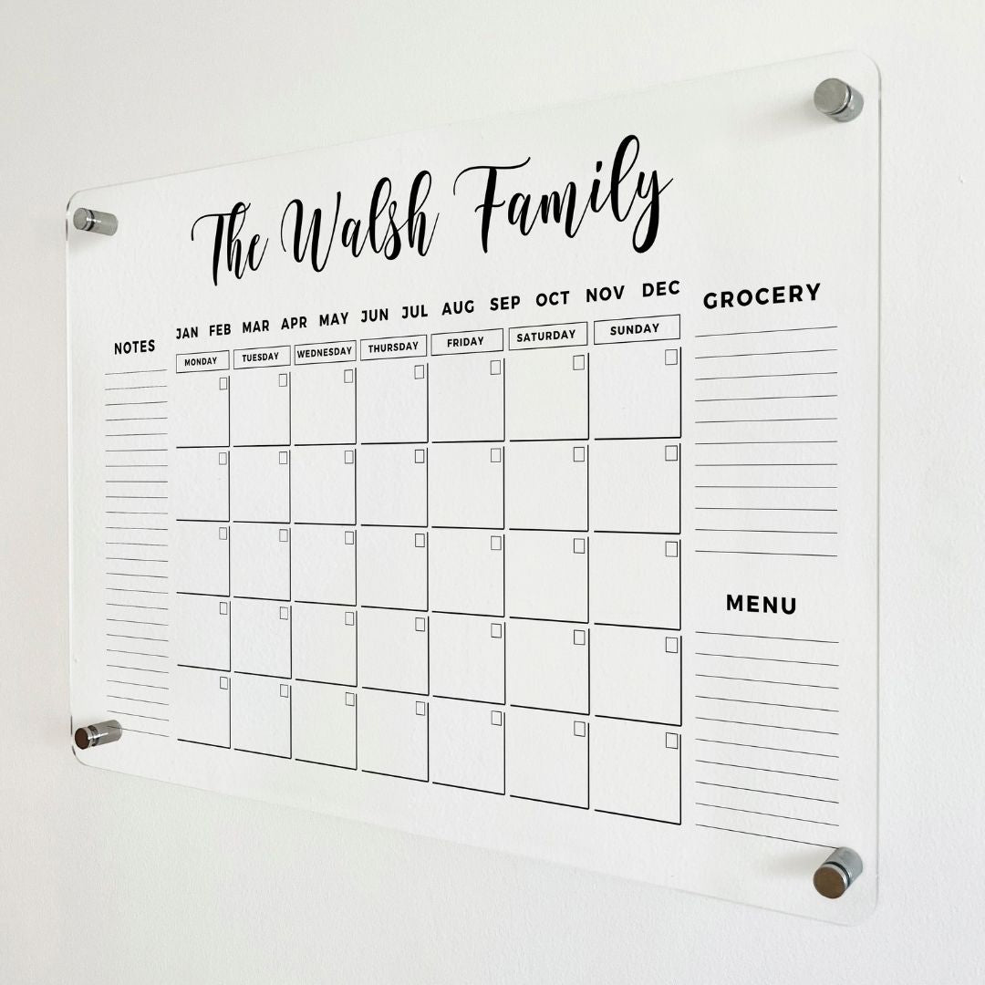 Angled close-up of a clear acrylic family calendar with a custom name heading, monthly grid, three side sections and silver wall mounts.