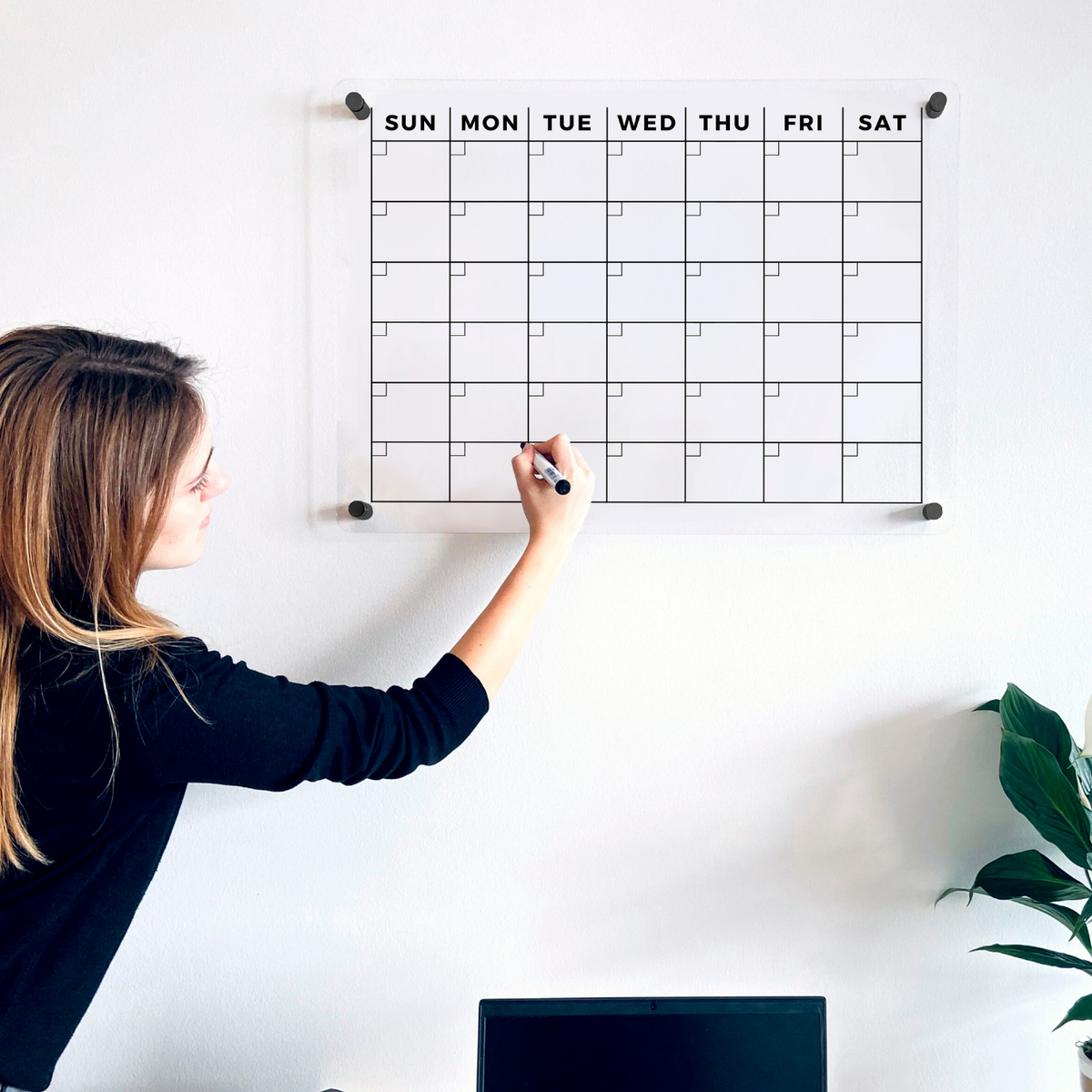 A person updating the date grid on a clear acrylic monthly wall planner above a desk.