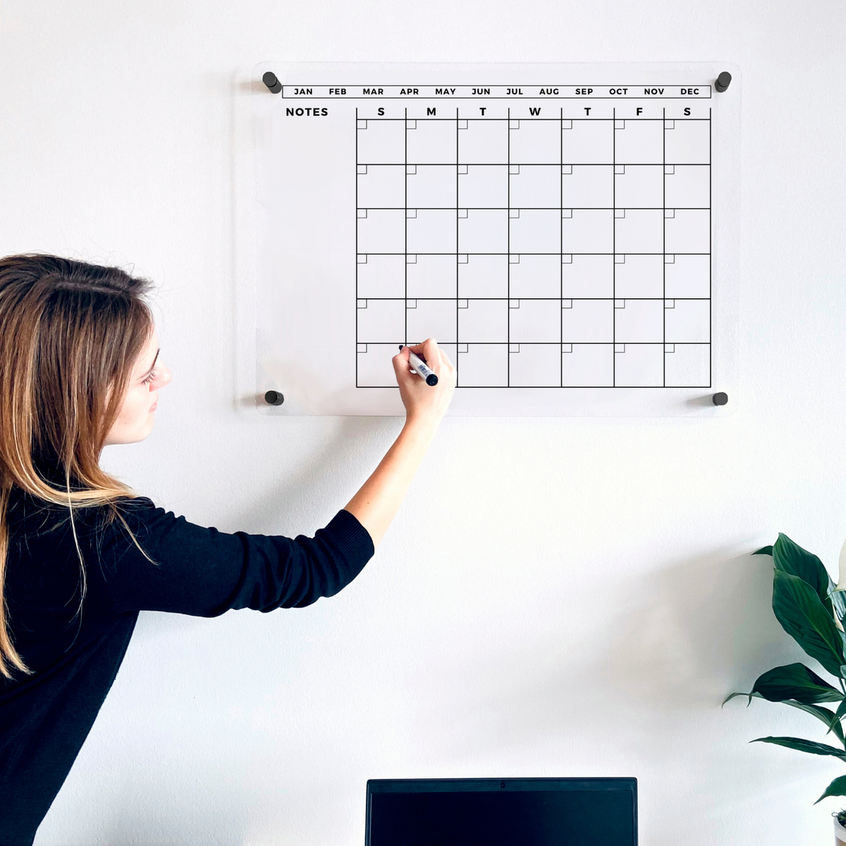 Woman writing on an acrylic monthly wall calendar with a large blank notes area on the left.