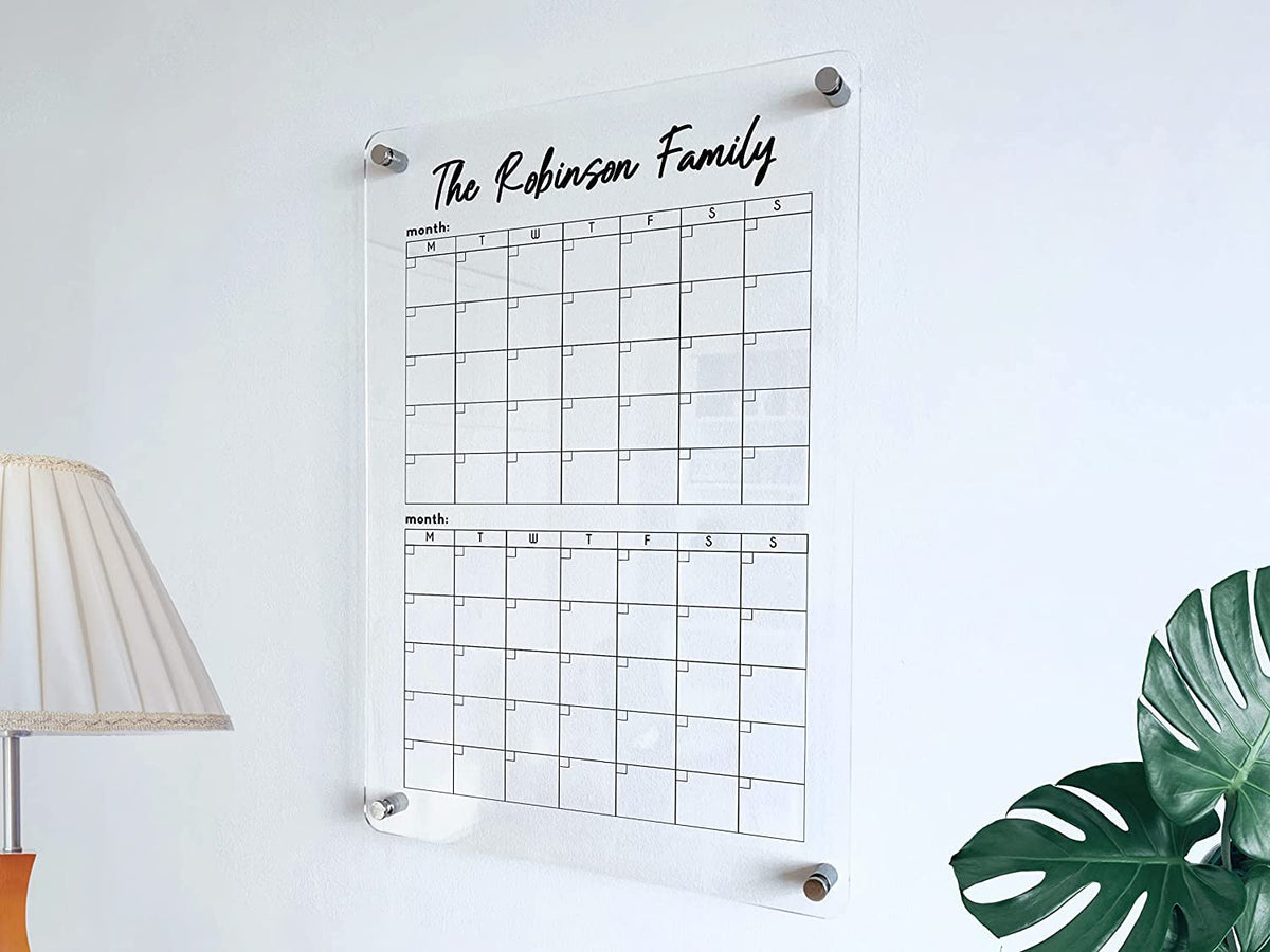 Vertical acrylic two-month family calendar displayed beside a lamp and plant, with two stacked date grids and gold mounts.