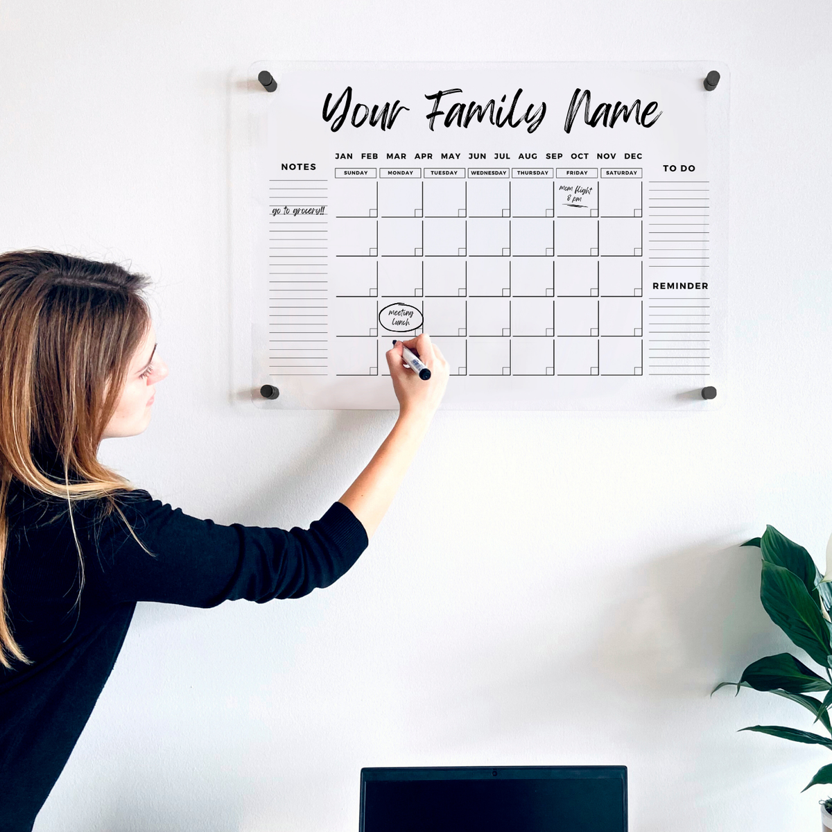Woman writing on an acrylic monthly wall calendar above a home office desk.