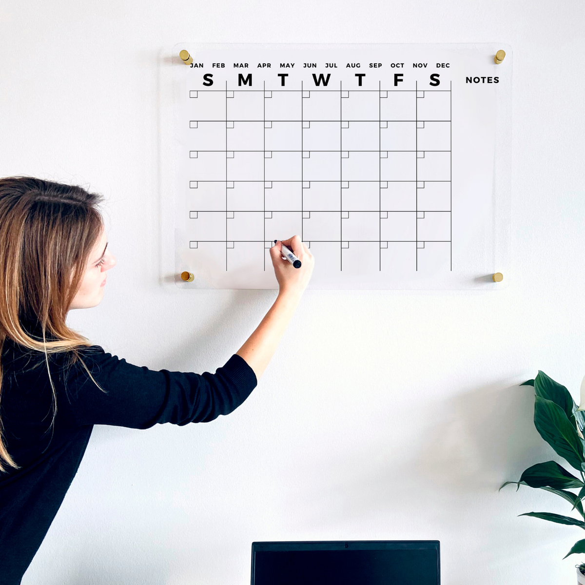 A person updating an acrylic monthly calendar with a right-hand Notes column above a desk.