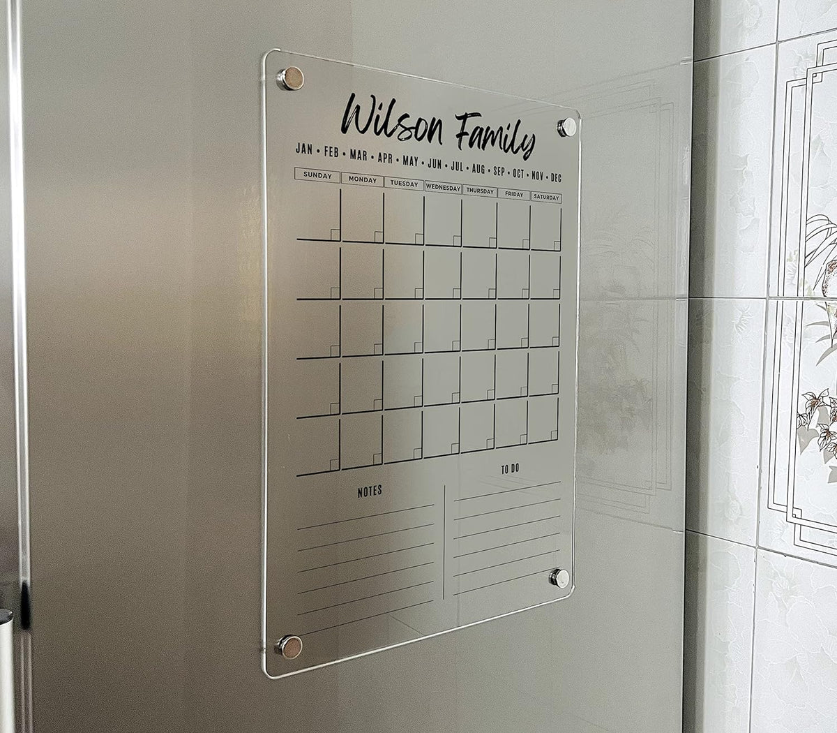 Angled view of a vertical magnetic acrylic calendar with two lined sections below its monthly grid on a refrigerator.
