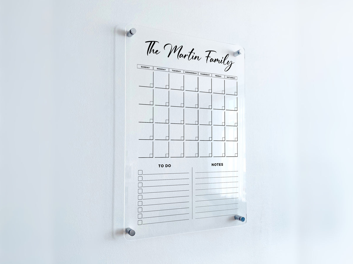 Angled view of a vertical acrylic monthly calendar with a custom family heading and two lined list sections beneath the date grid.