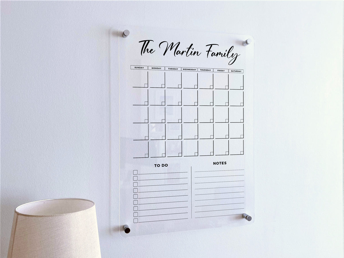 Vertical acrylic family calendar displayed above a lamp, with a monthly grid and two list sections below.